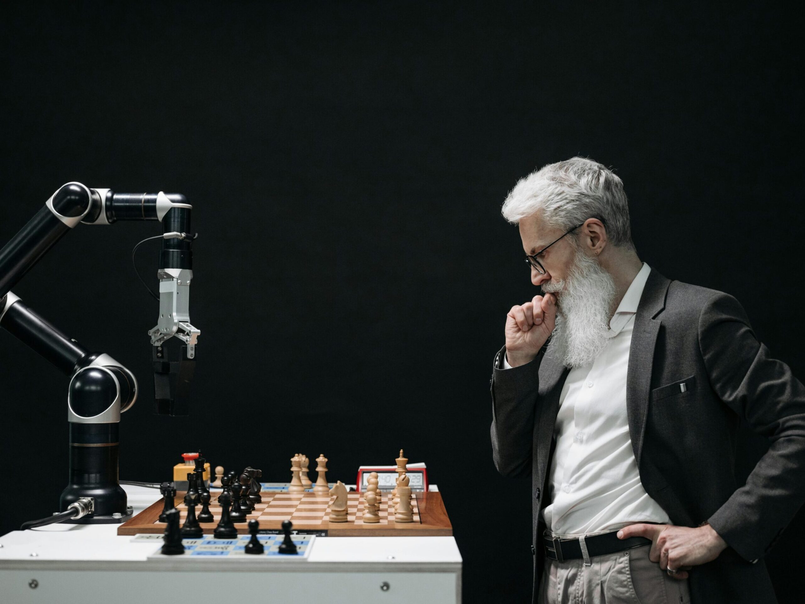 An elderly scientist contemplates a chess move against a robotic arm on a chessboard.