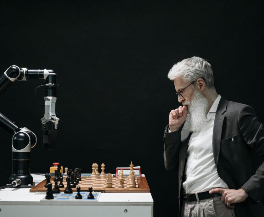 An elderly scientist contemplates a chess move against a robotic arm on a chessboard.